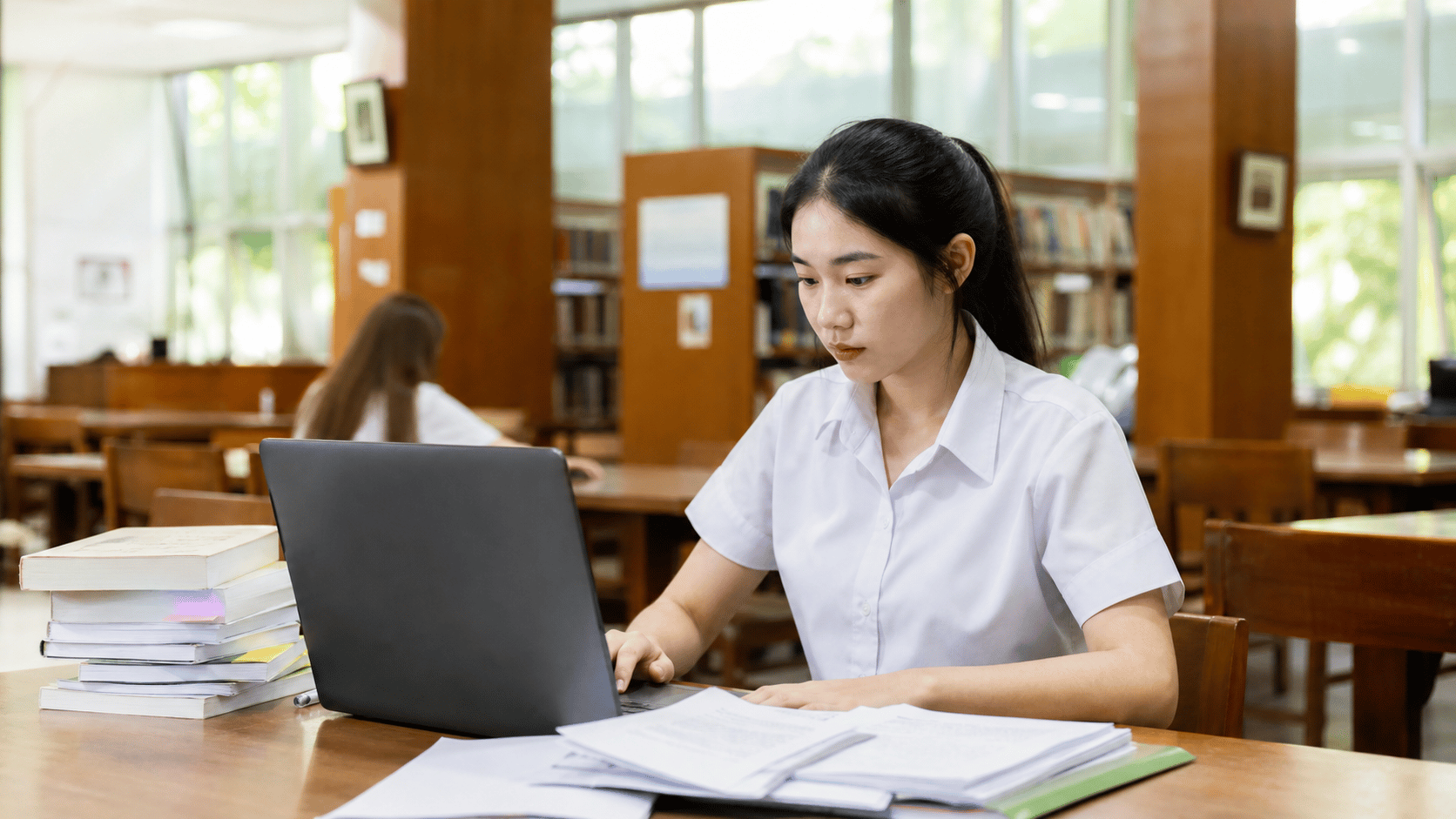 นักศึกษาไทยนั่งทำงานวิจัยด้วยแล็ปท็อปและเอกสารในห้องสมุดมหาวิทยาลัย